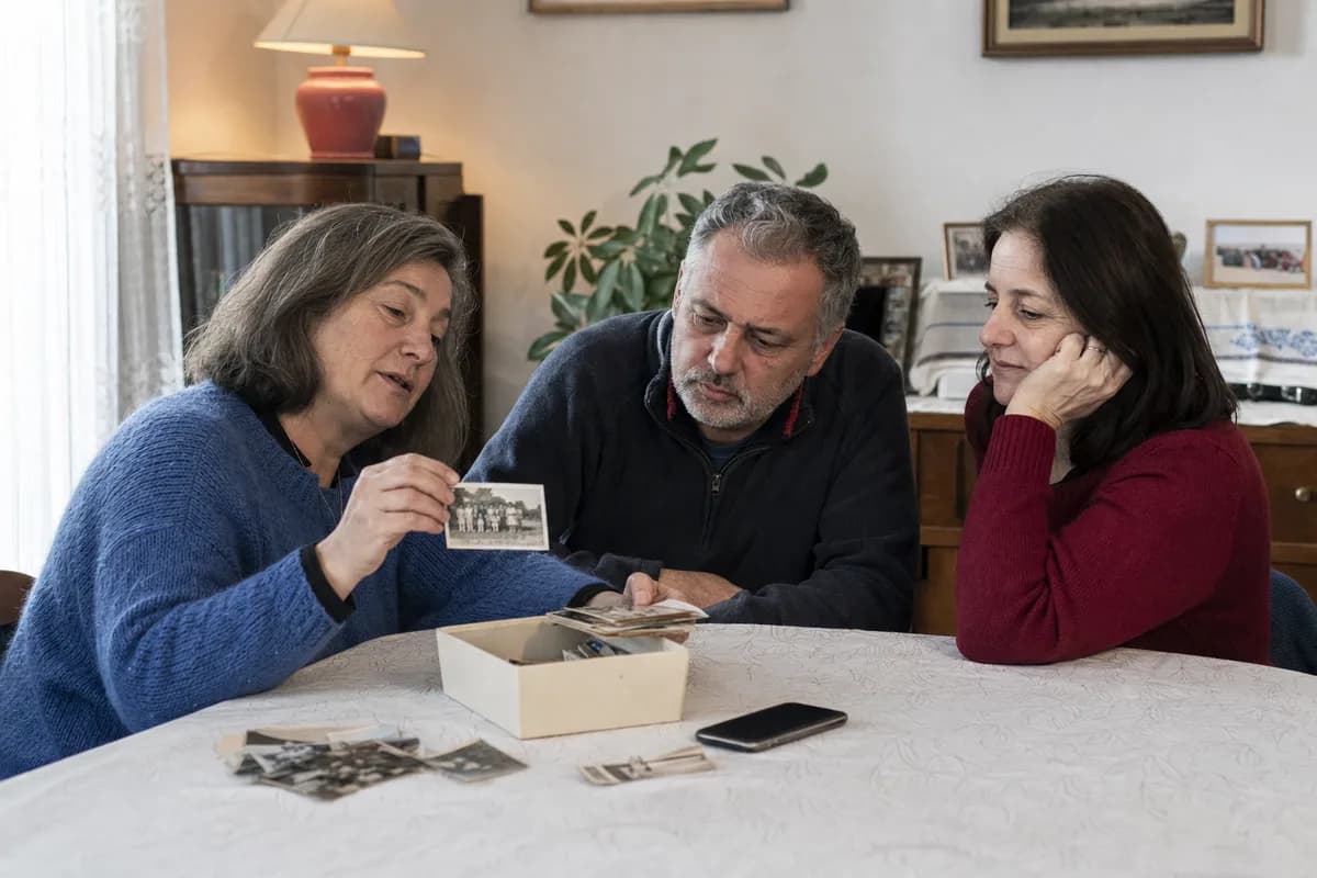 Three adult siblings sorting old family photographs around a dining table.
